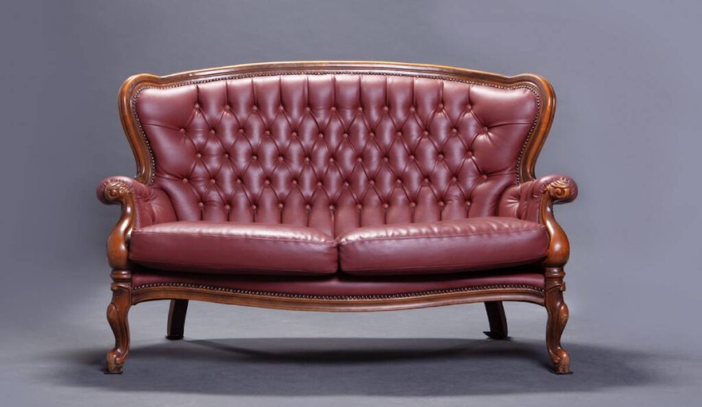 Elegant leather Chesterfield sofa with buttoned upholstery and carved wooden legs, set against a grey background.