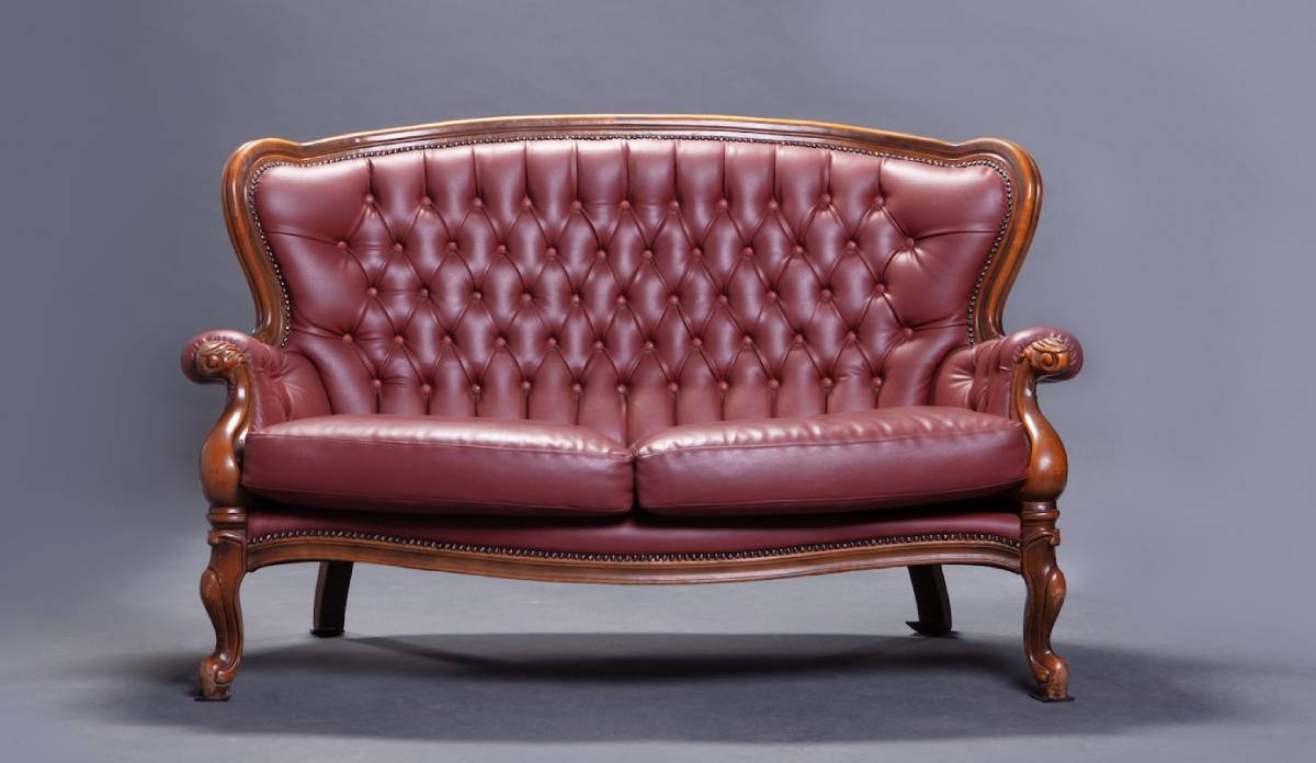 Elegant leather Chesterfield sofa with buttoned upholstery and carved wooden legs, set against a grey background.