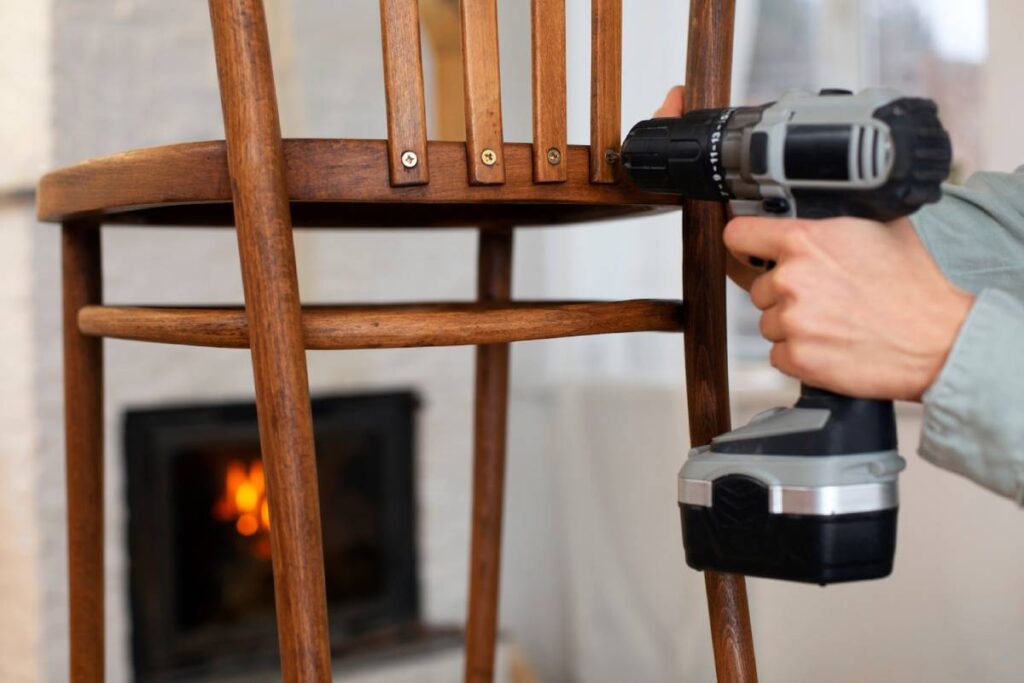 Person repairing a wooden chair with a cordless drill, near a fireplace.