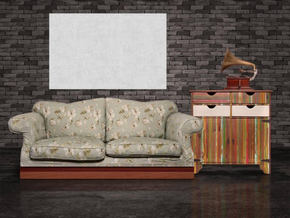 Floral sofa and a colourful cabinet with a gramophone on top, set against a dark brick wall with a blank white canvas. 