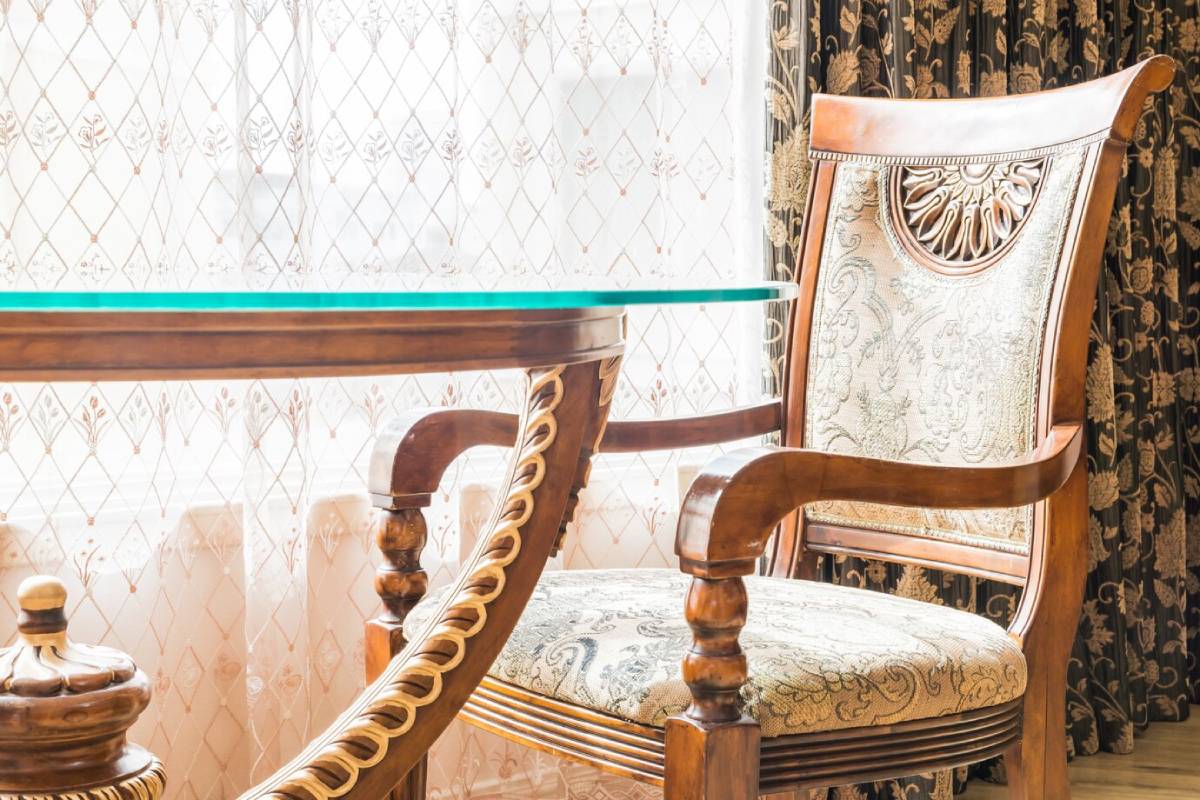  Elegant wooden chair with detailed fabric design next to a glass-top table, in a room with lace curtains.
