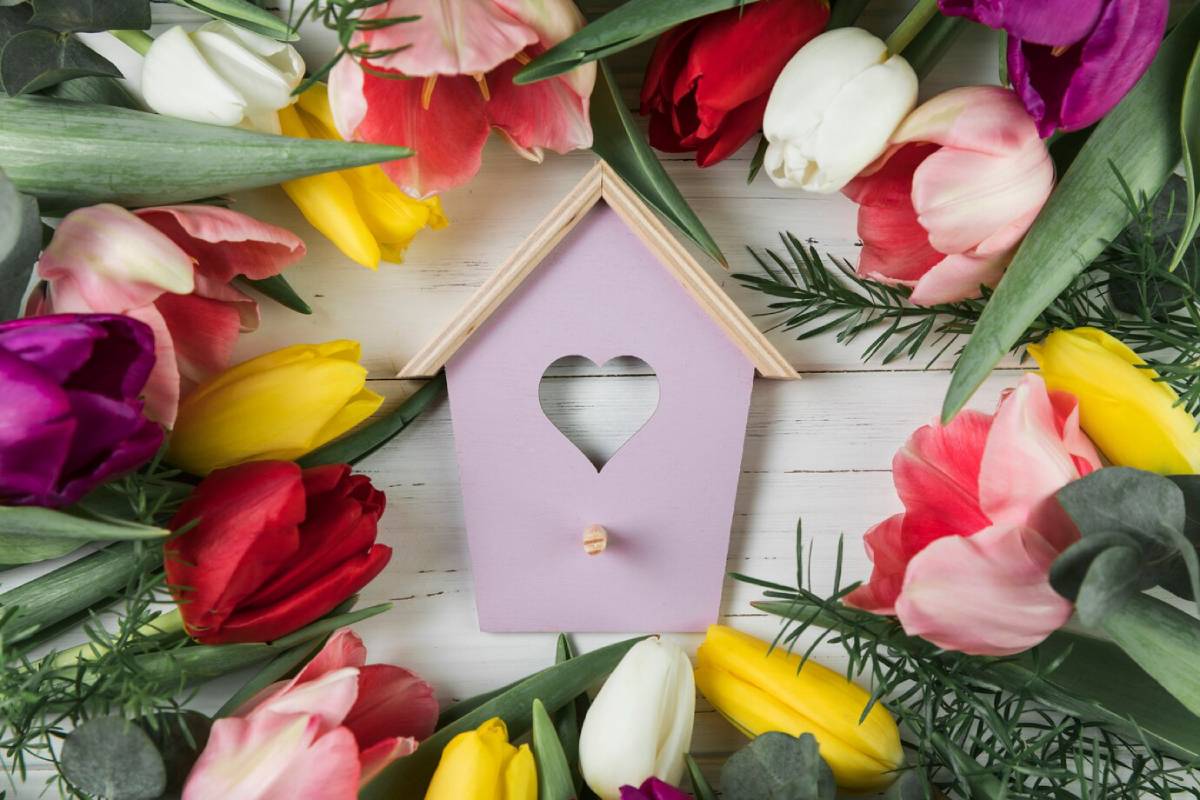 A small wooden birdhouse surrounded by colorful tulips on a white wooden background. 