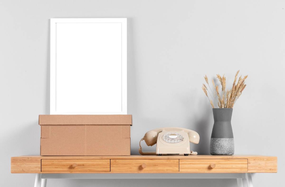 A wooden console table with a vintage telephone, cardboard boxes, and a vase with dried plants against a grey wall.