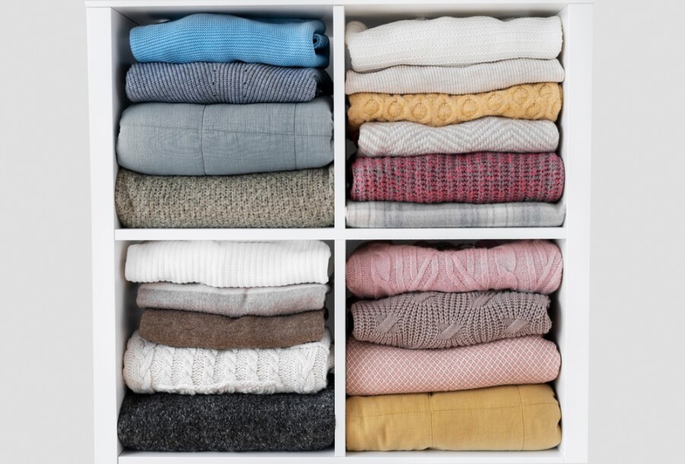  Four shelves of neatly folded jumpers in various colours and textures inside a white cupboard.