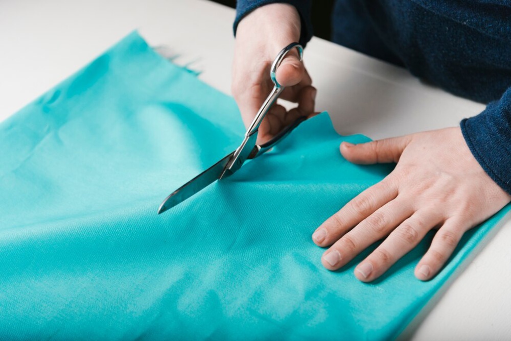 Person cutting a teal fabric with scissors on a white surface. 