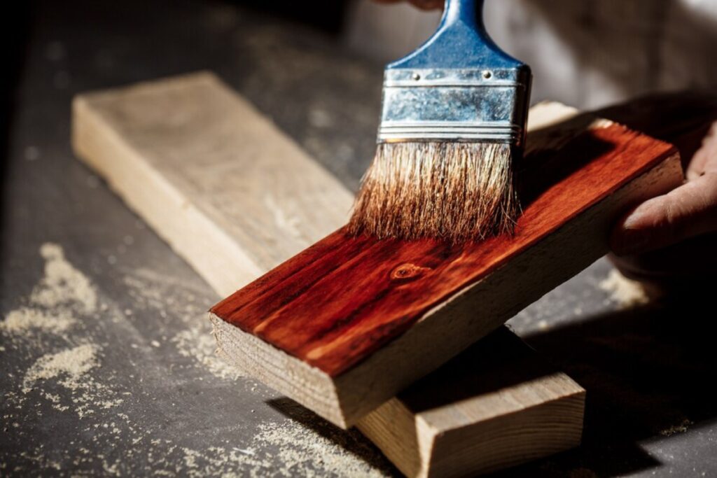 A person applying red paint to a wooden plank with a brush.