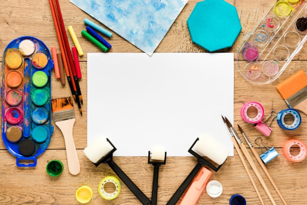 Assorted art supplies including paints, brushes, and a blank canvas on a wooden table. 