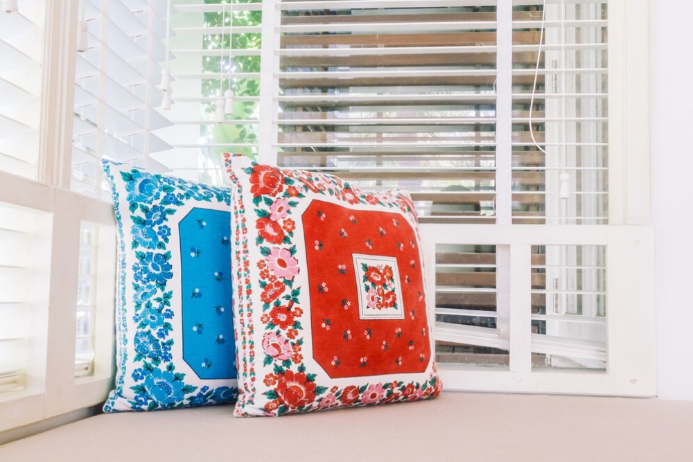 Two colourful floral cushions on a window seat, natural light filtering through white blinds. 