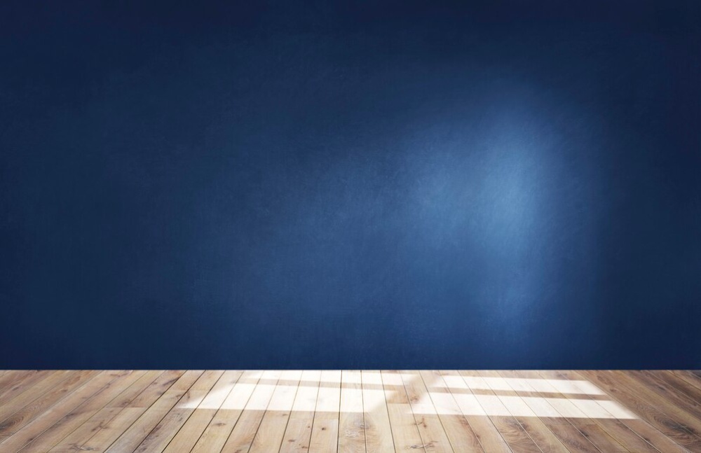 Empty room with a blue wall and wooden floor, illuminated by a beam of light from above. 
