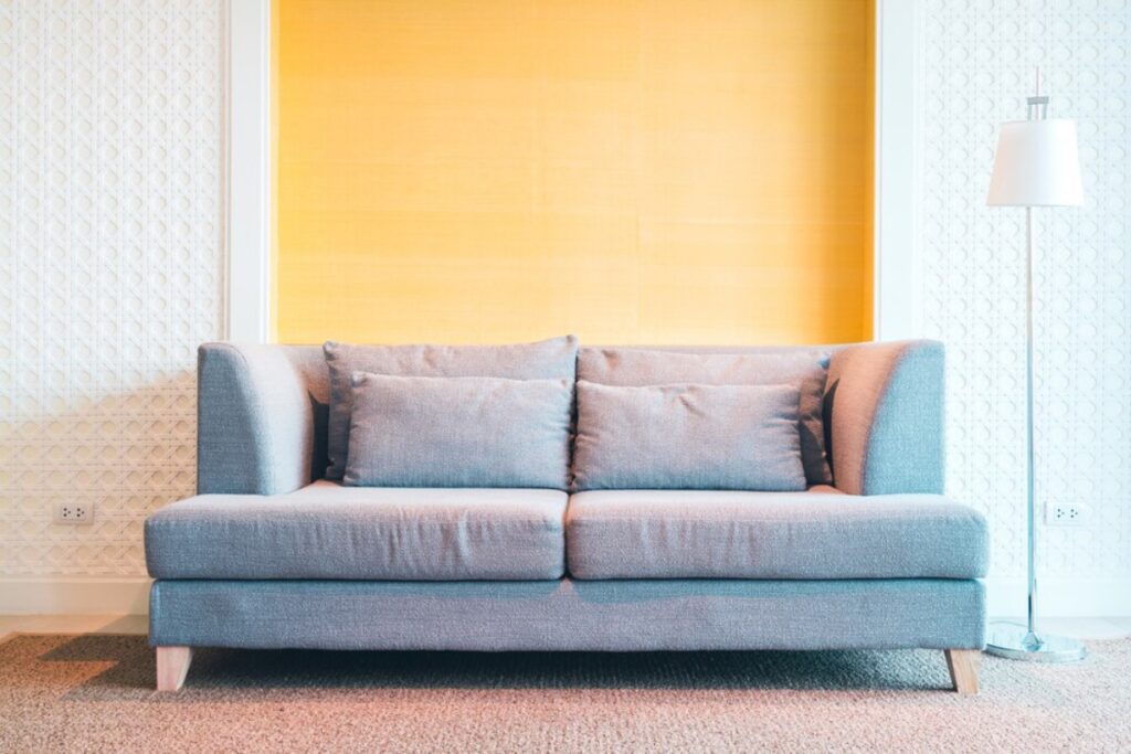A modern gray sofa with cushions in front of a vibrant yellow wall and a white lamp to the side.