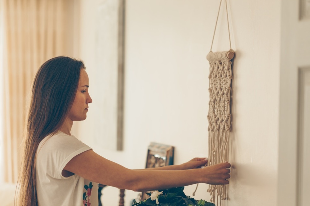 Woman in a white T-shirt adjusting a macramé wall hanging in a softly lit room.