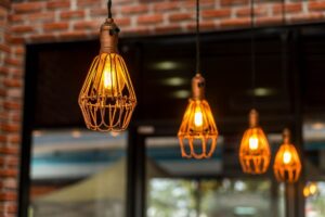 Warmly glowing Edison light bulbs in wire cages hanging from a ceiling, with a blurred brick wall and window background.