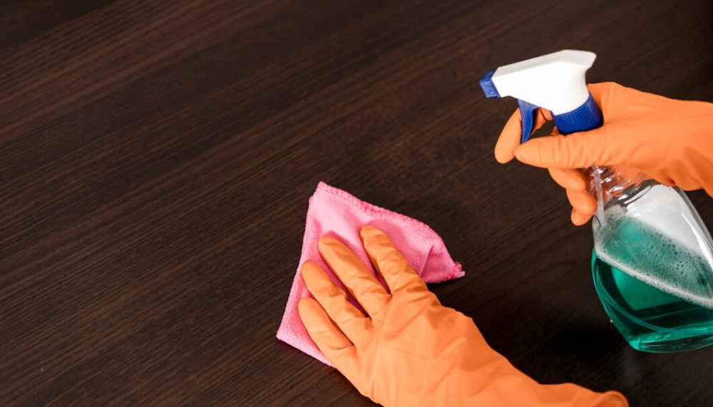  Hands in orange gloves cleaning a wooden surface using a pink cloth and a spray bottle with green liquid. 