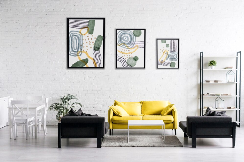 Modern living room with a yellow sofa, black armchairs, abstract wall art, and white brick walls. 