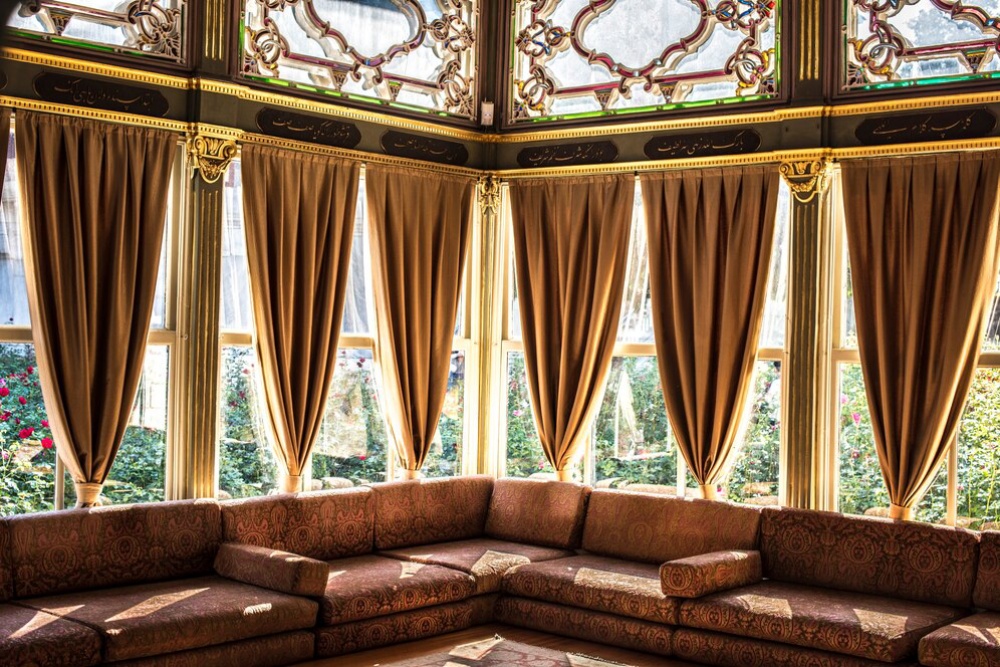  Opulent room with golden drapes, ornate ceiling, and floral-patterned sofas in front of sunlit windows overlooking a garden.