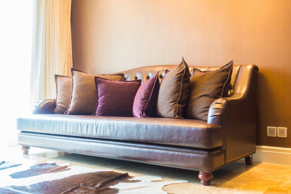 A luxurious brown leather sofa with many cushions, set against a textured beige wall in a warmly lit room. 