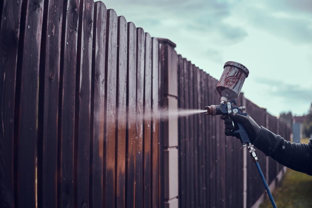  Person spraying dark stain on a wooden fence using a paint sprayer.