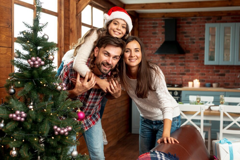  Family celebrating Christmas: a man giving a piggyback ride to a child in a Santa hat, with a woman beside them in a festive room.