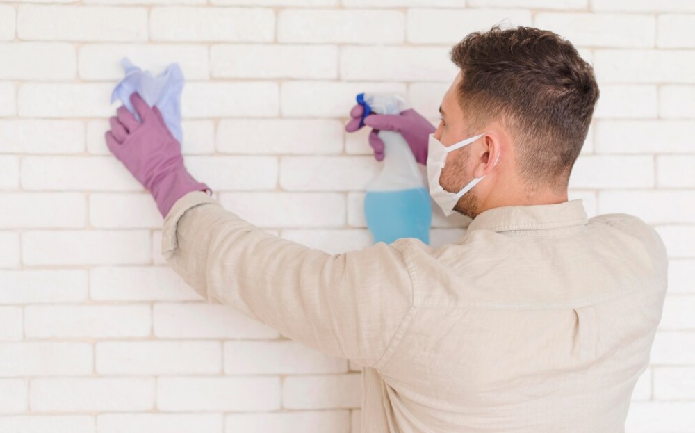 Man in mask and gloves cleaning a white brick wall with a cloth and spray bottle. 