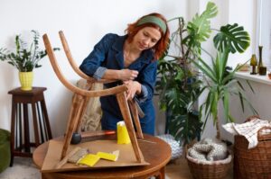 Woman refurbishes a wooden chair in a cosy, plant-filled room.