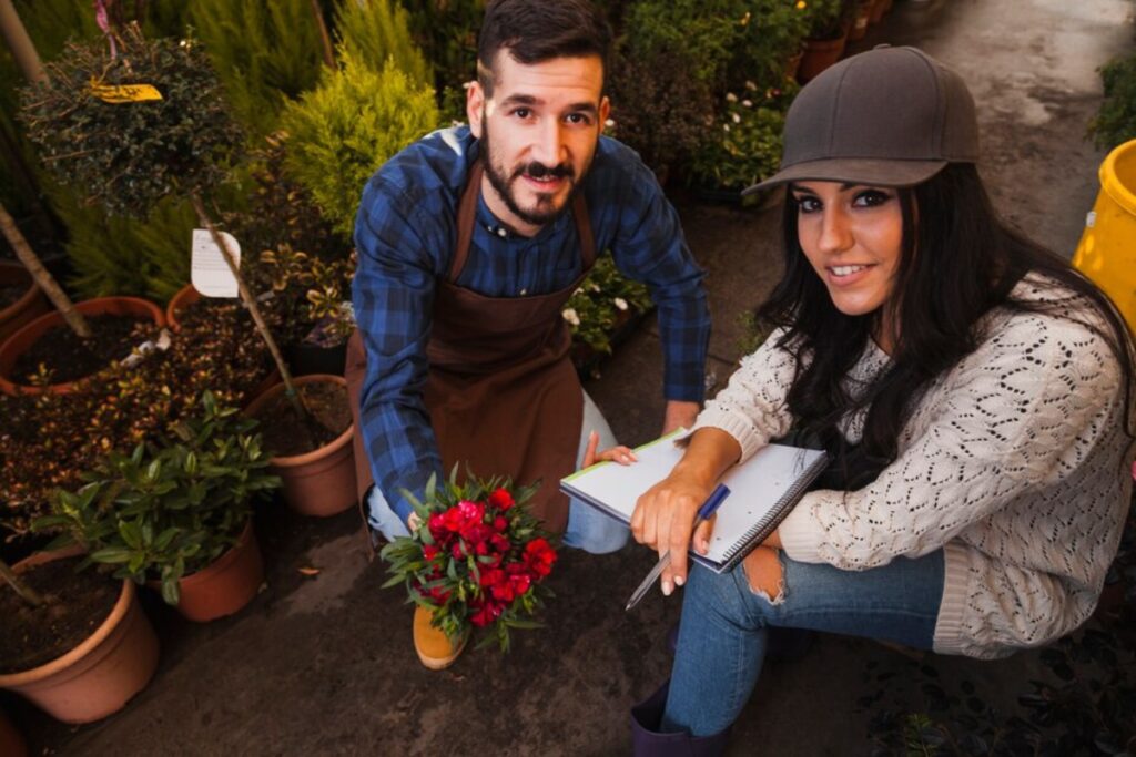 A man and woman discussing plants in a garden centre, surrounded by lush greenery and potted plants.