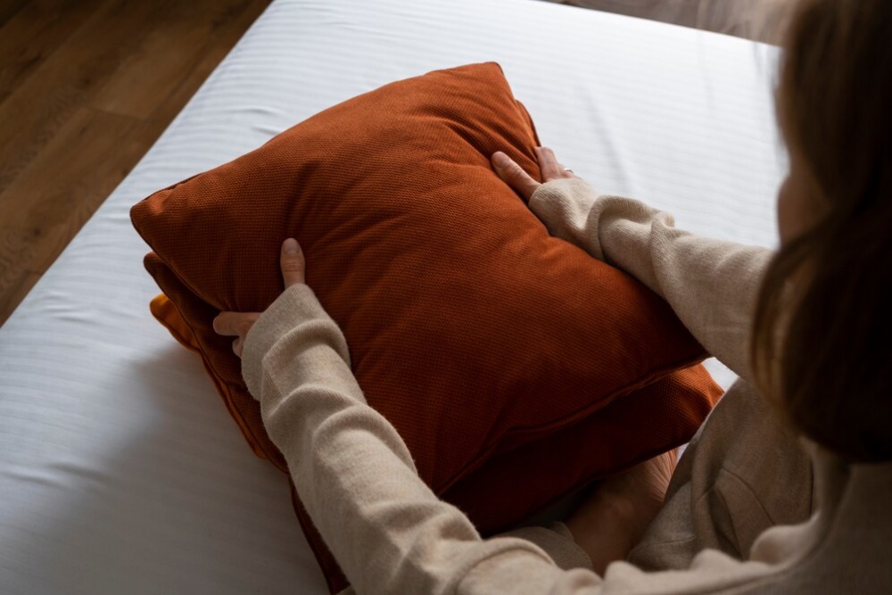  Person arranging a burnt orange pillow on a white bed, viewed from above.