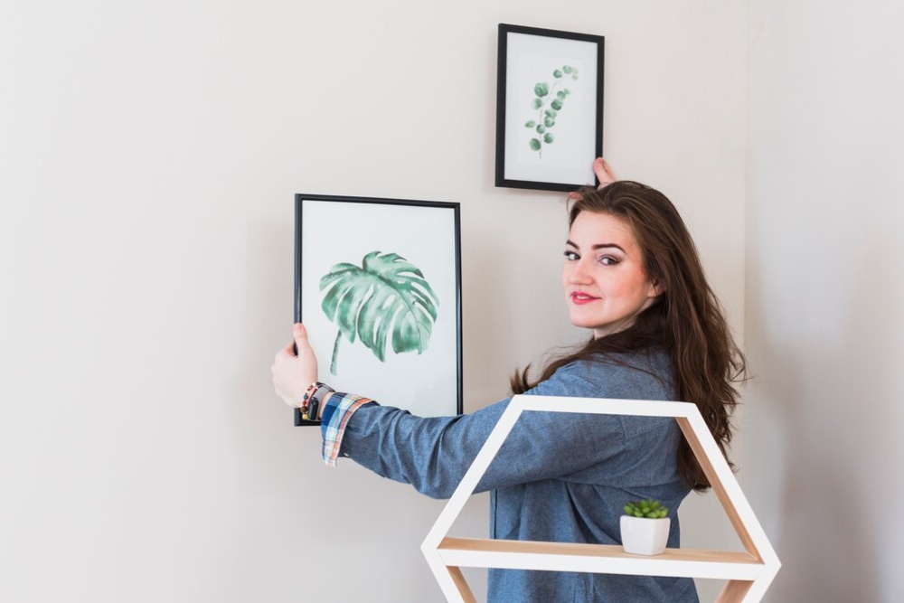  Woman hanging botanical artwork on a white wall in a bright room.