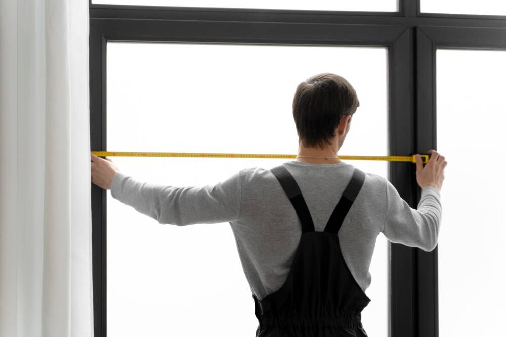 Man measuring a window with a tape measure from inside a room.