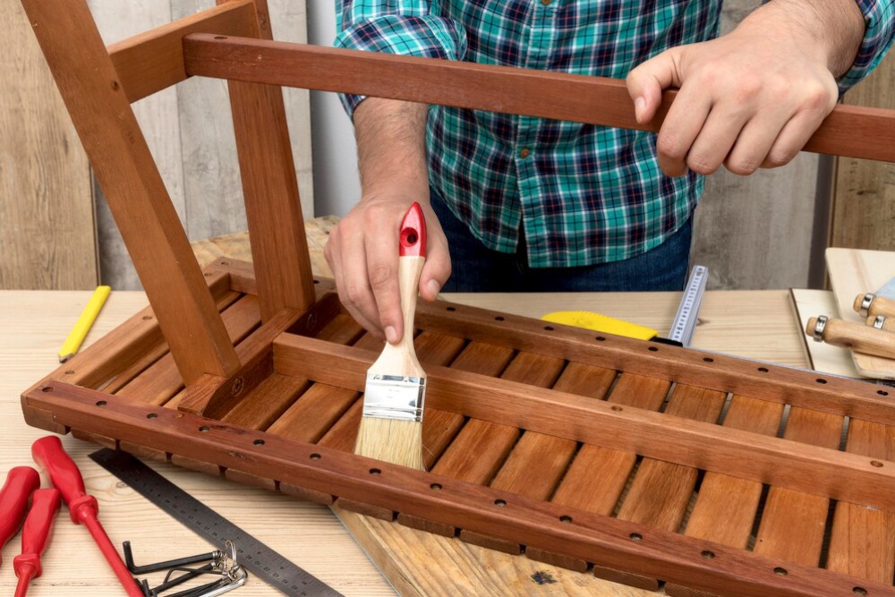  Man applying varnish to a wooden chair using a paintbrush, surrounded by tools on a workbench.