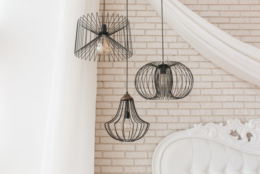  Three decorative metal pendant lights hanging above a white ornate bed, against a brick wall backdrop.