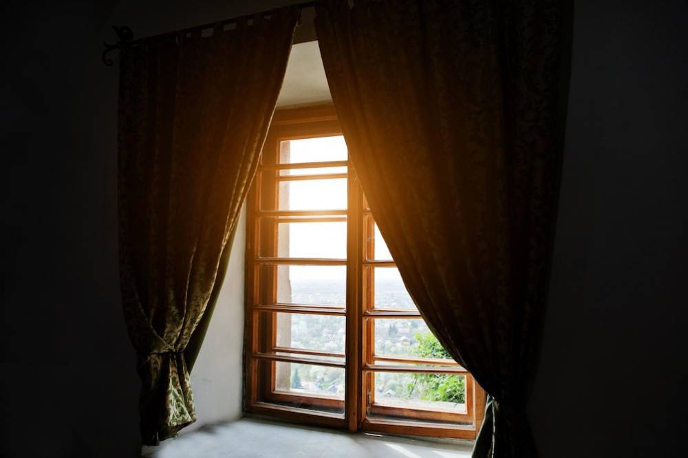 Sunlight streams through an open window with ornate curtains in a room, highlighting the peaceful view outside.