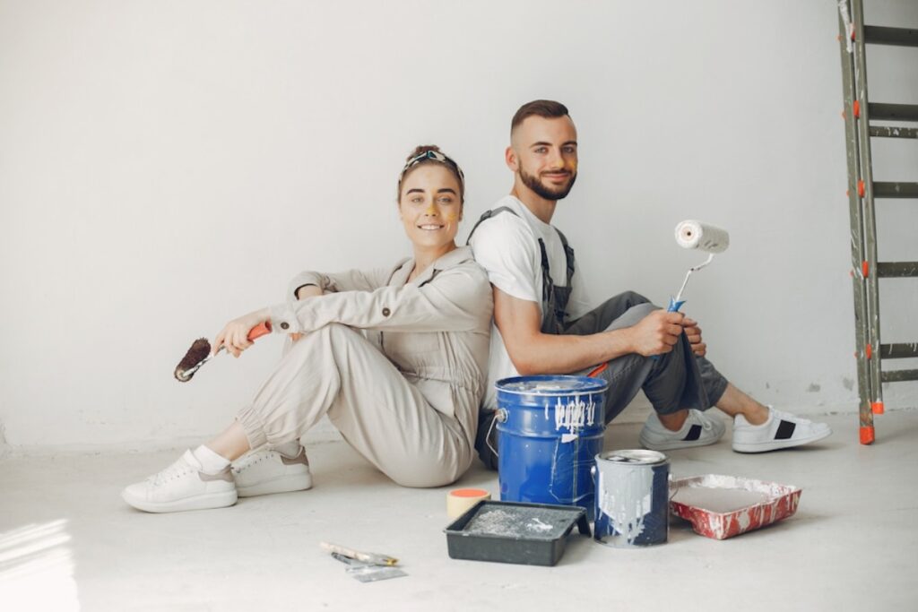 Two smiling people in overalls sitting with paint supplies in a white room.