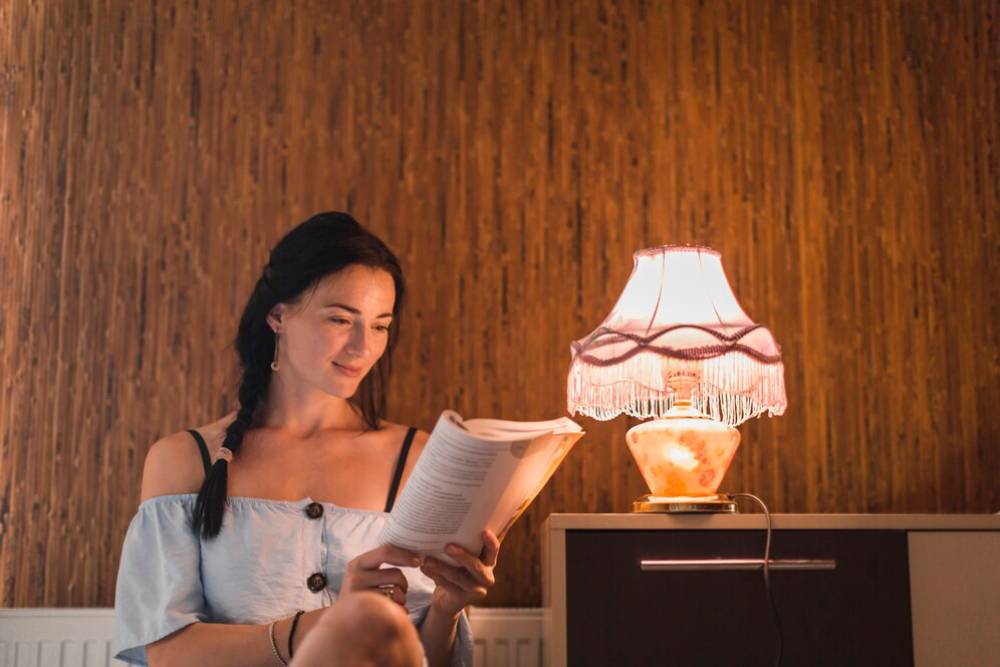  Woman reading a book by a table lamp in a warmly lit room with rustic wooden wall.